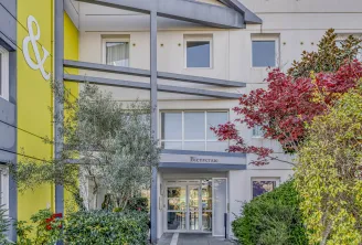 B&B Hotel Bordeaux Bruges entrance with 'Bienvenue' sign, modern white and yellow facade with red flowering plants and climbing ivy