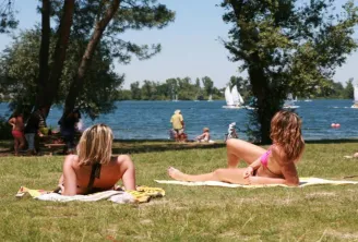 Lake Beach in Bordeaux: visitors relax on the grass overlooking the lake with sailboats. Trees frame the scene while other swimmers enjoy the water in the background.