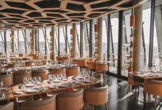 The 7 panoramic restaurant at La Cité du Vin in Bordeaux featuring elegant light wood tables, caramel-colored chairs, wine glasses, modern geometric ceiling, and floor-to-ceiling windows overlooking the city