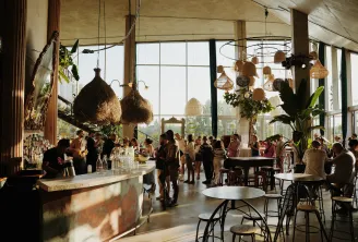 Modern bar interior in Bordeaux featuring floor-to-ceiling windows, bohemian hanging basket décor, marble counter, relaxing guests, and city views in the background.