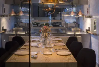 Modern dining room with light wooden table, black chairs, suspended golden pendant lights, glass partitions and open kitchen visible in the background, located at Cent33 in Bordeaux.