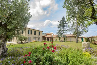Bordeaux wine estate Domaine des Perchés featuring stone buildings, flowering garden with red roses and blue flowers, ancient olive trees and tree-lined pathways under cloudy sky.