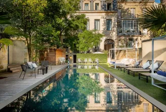 A rectangular reflection pool surrounded by lush green gardens, lounge chairs, and relaxation areas overlooking the elegant classical façade of Palais Gallien Hôtel & Spa in Bordeaux
