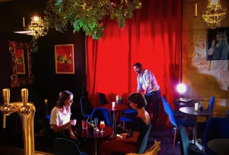 Interior of Théâtre des Chartrons in Bordeaux featuring three people in an elegant bar with red walls, blue furniture, sophisticated lighting and plant decoration.