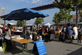 Lively market in Lormont with visitors under blue umbrellas, product stalls, and the stone bridge visible in the background under clear sky.