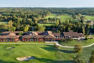 Aerial view of a luxury hotel complex with red-orange facades and brown roofs, surrounded by a lush golf course with bunkers, water features, and forests near Bordeaux.