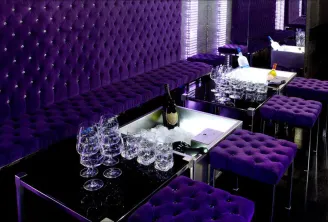 VIP lounge at Boomboom Club in Bordeaux featuring purple tufted banquettes, crystal-studded padded walls, black tables, champagne glasses and bottle in ice bucket, with ambient purple LED lighting.