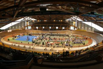 Panoramic view of Bordeaux velodrome featuring its red clay oval cycling track, packed spectator stands, and distinctive wooden and glass architecture with an arched roof.