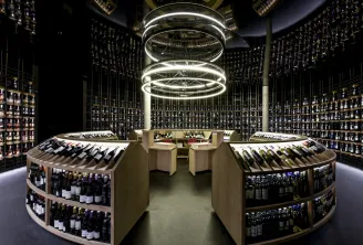 Modern wine cellar interior with curved wooden shelving displaying bottles, black walls with storage racks, and a spiral-shaped ceiling light fixture.
