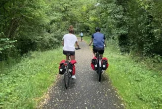 Two cyclists riding red bicycles on a gravel bike path surrounded by green vegetation in Bordeaux, with other cyclists visible in the background.