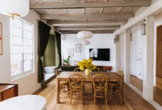 Modern loft interior featuring a wooden dining table with rattan chairs, yellow flowers, exposed beams, and large windows overlooking Bordeaux