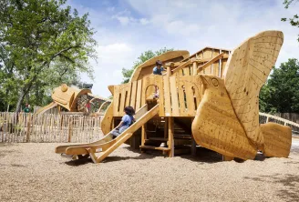 Wooden boat-shaped playground with slide, children playing, surrounded by wooden fence, at Forêt du Bourgailh in Bordeaux.