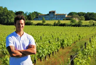 A smiling man in a white t-shirt standing in front of Bordeaux vineyards with a historic estate in the background