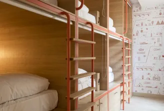 Modern dormitory room with red metal-framed bunk beds, wooden shelves, white bedding, integrated LED lighting, and travel-themed wallpaper
