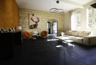 Contemporary interior of Clos des Jacobins in Bordeaux featuring a spacious living area with exposed stone walls, modern furniture in neutral and orange tones, minimalist décor with a deer portrait, large arc lamp and large windows.