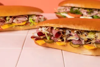 Three sandwiches filled with cold cuts, melted cheese, pickled onions and lettuce on golden bread rolls, displayed on a white surface with coral background.