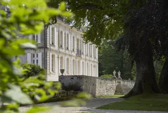 View of Château de La Dauphine in Bordeaux featuring its white neoclassical facade with columns, surrounded by lush greenery and ancient trees, accessed by stone steps.