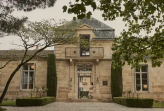 Classical stone facade of a Bordeaux castle featuring a central ornate doorway, wrought iron balcony, flanked cypress trees, and manicured garden landscaping