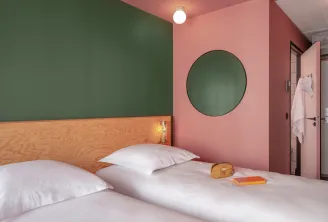 Modern bedroom at Eklo Bordeaux featuring a double bed with light wood headboard, sage green and coral pink walls, round green mirror, white ceiling light, and bathroom door.