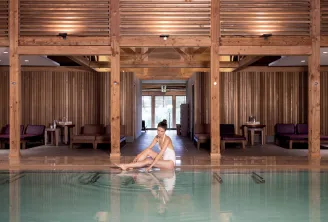 A woman sitting at the edge of an indoor swimming pool in a spa with massive wooden architecture and relaxation areas in the background.