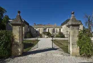 Entrance gates of Château Picque Caillou in Bordeaux, featuring ornate stone pillars with spherical finials, gravel driveway, and classical mansion facade against clear blue sky.