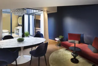 Luxury suite at Hotel Cardinal Bordeaux featuring a modern living space with red sofa, white marble dining table, black chairs, a spa-like bathroom with walk-in shower, and navy and gold tones