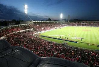 Aerial view of the illuminated Chaban-Delmas Stadium in Bordeaux during evening, filled with spectators in red and white attire, rugby pitch in the foreground with crowded stands.