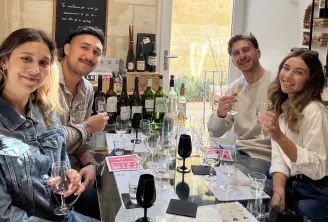 Four smiling young adults at a wine tasting in Bordeaux, seated around a table with multiple wine bottles and glasses.