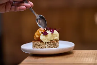 Gourmet plated dessert featuring an almond cake base, light mousse, golden sponge cake and purple edible flowers with a spoonful of sauce being drizzled on top