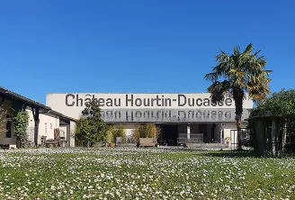 Facade of Château Hourtin-Ducasse in Bordeaux with entrance sign, surrounded by flowering garden and palm tree against blue sky