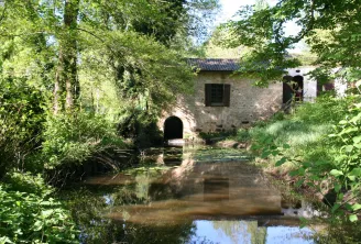 Historic stone building with an arched entrance and windows surrounded by dense green vegetation, crossed by a calm waterway in the Eau Bourde valley near Bordeaux.