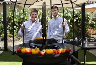 Two chefs in white uniforms pose in front of a grill with fresh tomatoes at Lafaurie Terrace in Bordeaux, surrounded by white and pink flowers.