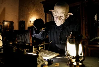 Fantastical creature in black costume and makeup in a gothic Bordeaux setting, surrounded by glowing lanterns and antique objects.