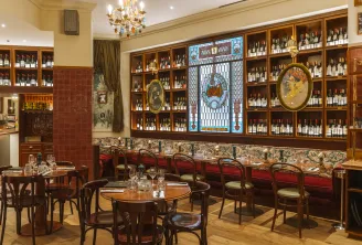 Elegant interior of Brasserie Vinatier in Bordeaux featuring extensive wine bottle displays in wooden cabinets, round wooden tables, classic bentwood chairs, red velvet banquette seating, decorative stained glass windows, and a golden crystal chandelier.