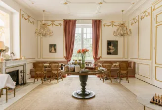 Elegant neoclassical drawing room at Château Léognan in Bordeaux, featuring cream walls, gilt wood paneling, two ornate chandeliers, red curtains, round central table, fireplace, antique portraits, and wicker furniture.