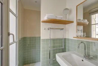Modern bathroom at Badon Boutique Hôtel in Bordeaux featuring mint green tile, white sink, round mirror, wooden shelves, white towels and bathtub with glass partition.