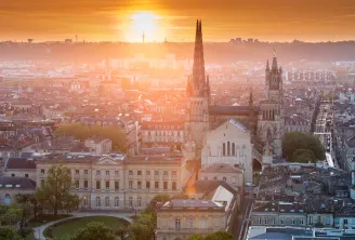 Aerial view of Bordeaux at sunset featuring the Basilica of Saint-Michael and its two Gothic spires in the foreground, overlooking the city's classical architecture and historic districts.