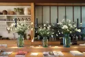 Wooden table featuring three glass vases with white and yellow daisies, black hanging lights above, modern kitchen in the background, magazines on the table, bright and elegant interior.