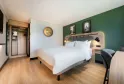 Modern hotel room at Campanile Nature Bordeaux Lac featuring a double bed with white linens, green accent wall, workspace, open wardrobe and contemporary bathroom