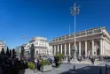 The Grand Theatre of Bordeaux, a neoclassical building with majestic white columns, ornamental façade and clock, overlooking a bustling public square under clear blue sky.