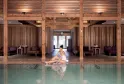 A woman sitting by the edge of an indoor swimming pool with turquoise waters in a spa with massive wooden structures, featuring relaxation cabins in the background at Sources de Caudalie in Bordeaux.