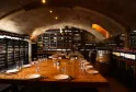 Arched underground wine cellar featuring a wooden tasting table, warm ambient lighting, and shelves stocked with Bordeaux wine bottles