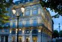 Illuminated facade of the Hôtel de Sèze in Bordeaux, a historic Haussmann-style building featuring columns, wrought-iron balconies and mansard roof, photographed at dusk with a street lamp in the foreground.