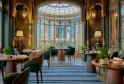 Elegant restaurant dining room featuring ornamental glass windows, round wooden tables, blue chairs, golden wall lamps, and a geometric glass dome ceiling with colorful panes.