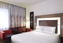 Modern hotel room at Novotel Bordeaux Lac featuring a king-size bed with white linens, a seating area with red armchair, gray and red curtains, and contemporary decor.