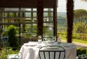 Fine dining table set with white tablecloth, glassware, and place settings on a terrace overlooking Bordeaux vineyards and valley landscape, framed by stone columns and large windows.