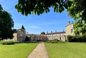 Courtyard of Château de Portets near Bordeaux featuring stone buildings with a conical tower, central pathway, and manicured green lawns against a clear blue sky.