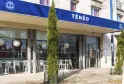 Facade of Ténéo Aparthotel in Talence Centre, Bordeaux, featuring modern large windows, blue and white signage, outdoor terrace with white chairs and green topiary plants.