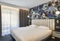 Modern hotel room featuring a double bed with white linens, beige upholstered headboard, black accent wall with blue and white floral mural, brown curtains, window with sheer drapes and round pendant lights.