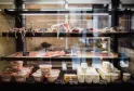 Delicatessen display case featuring various types of cured meats, sliced charcuterie, and aged cheeses arranged on three illuminated shelves with handwritten price labels.